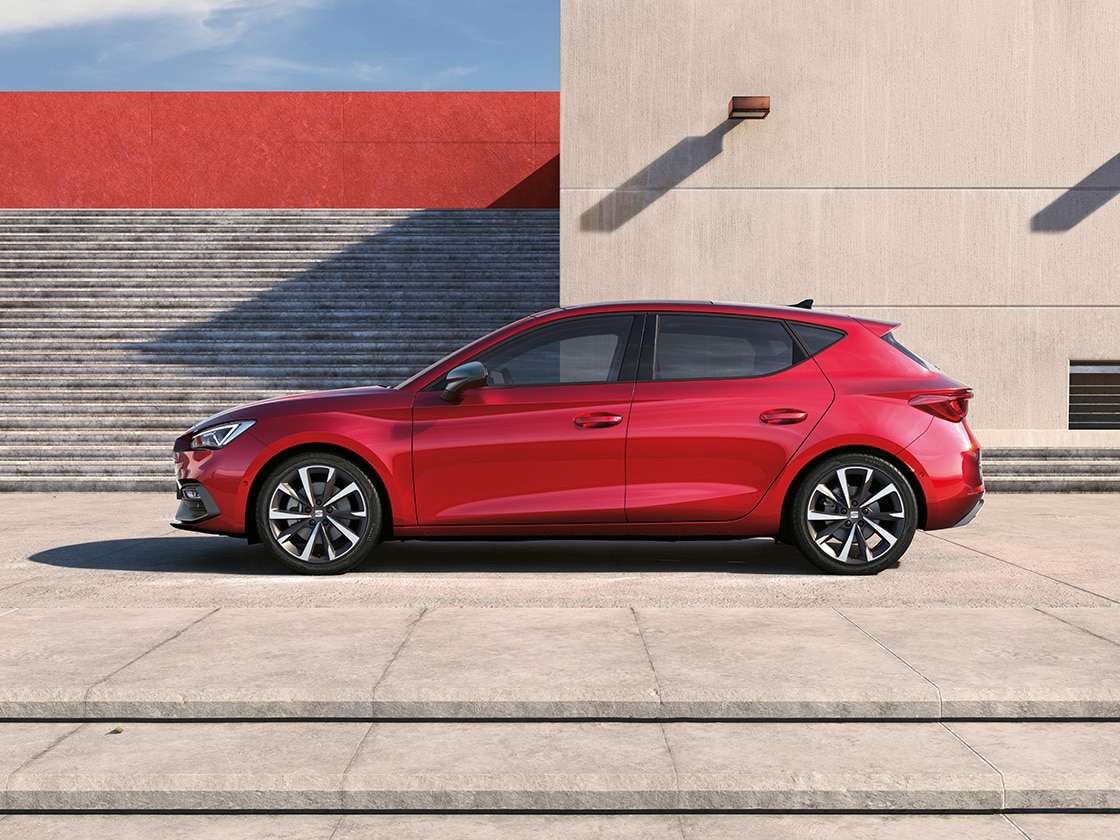 Side view of the SEAT Leon NF 5D in desire red parked outdoors, in a sunny spot with a building and stairs behind.